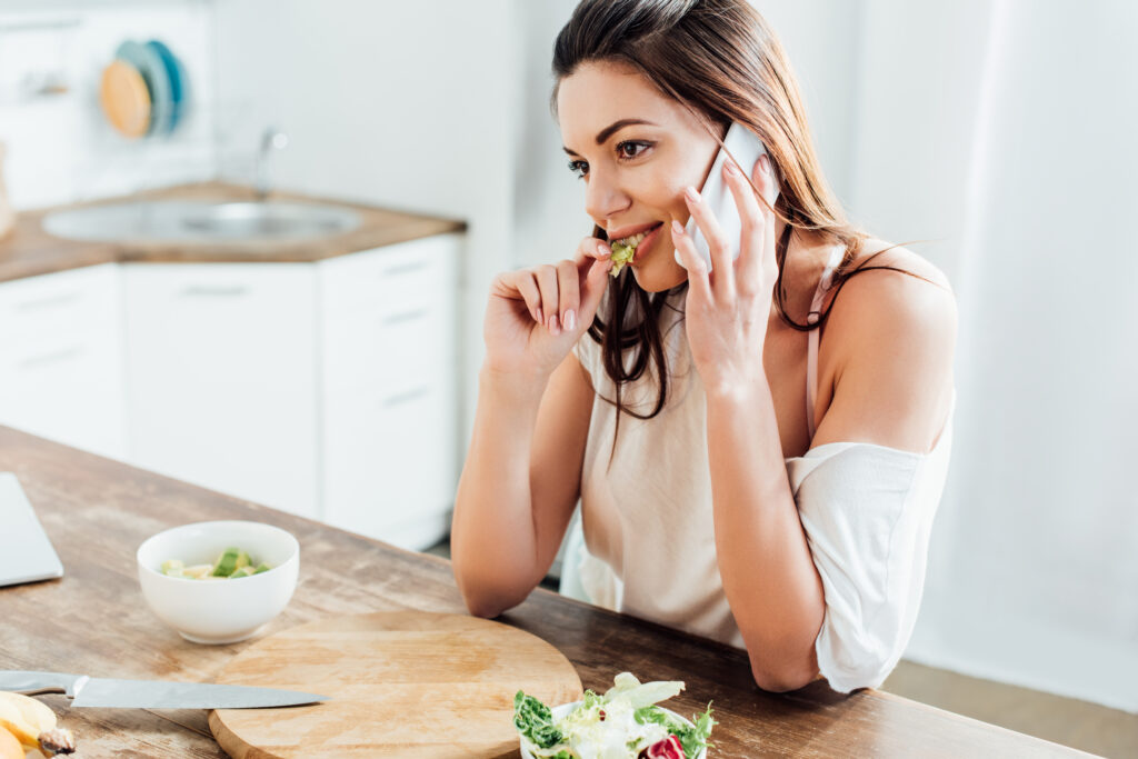 Jeune femme assise dans sa cuisine, au téléphone tout en mangeant de la salade. 