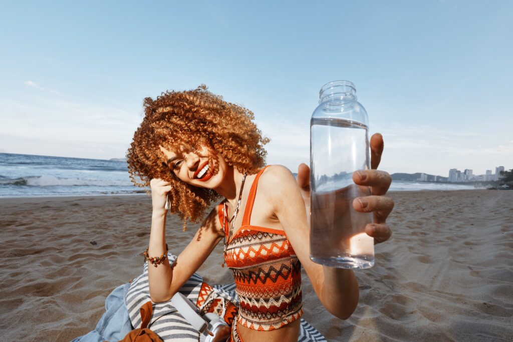 Jeune femme très souriante, aux cheveux frisés, assise sur une plage et brandissant une bouteille d'eau. 